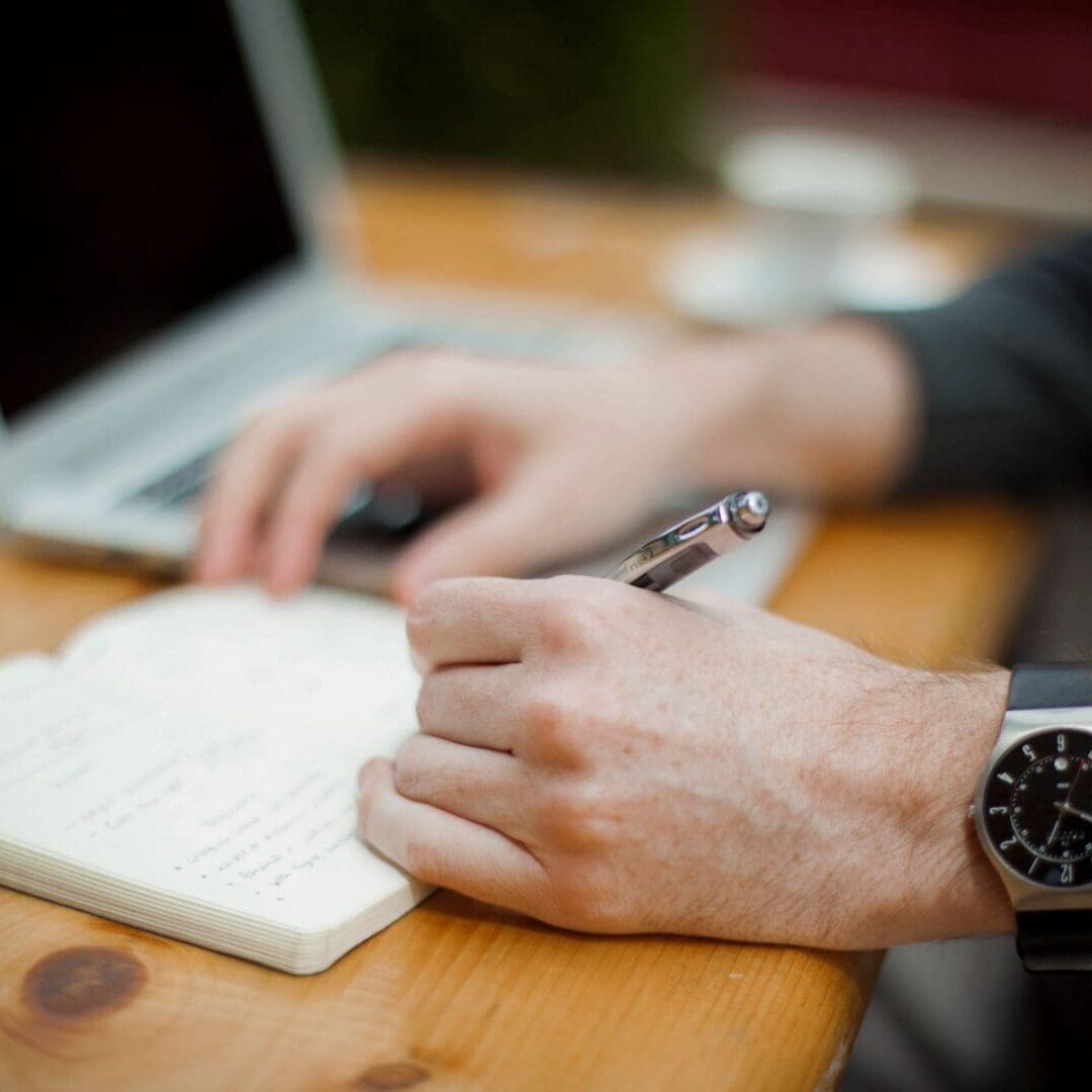 A person is writing on paper with a laptop.